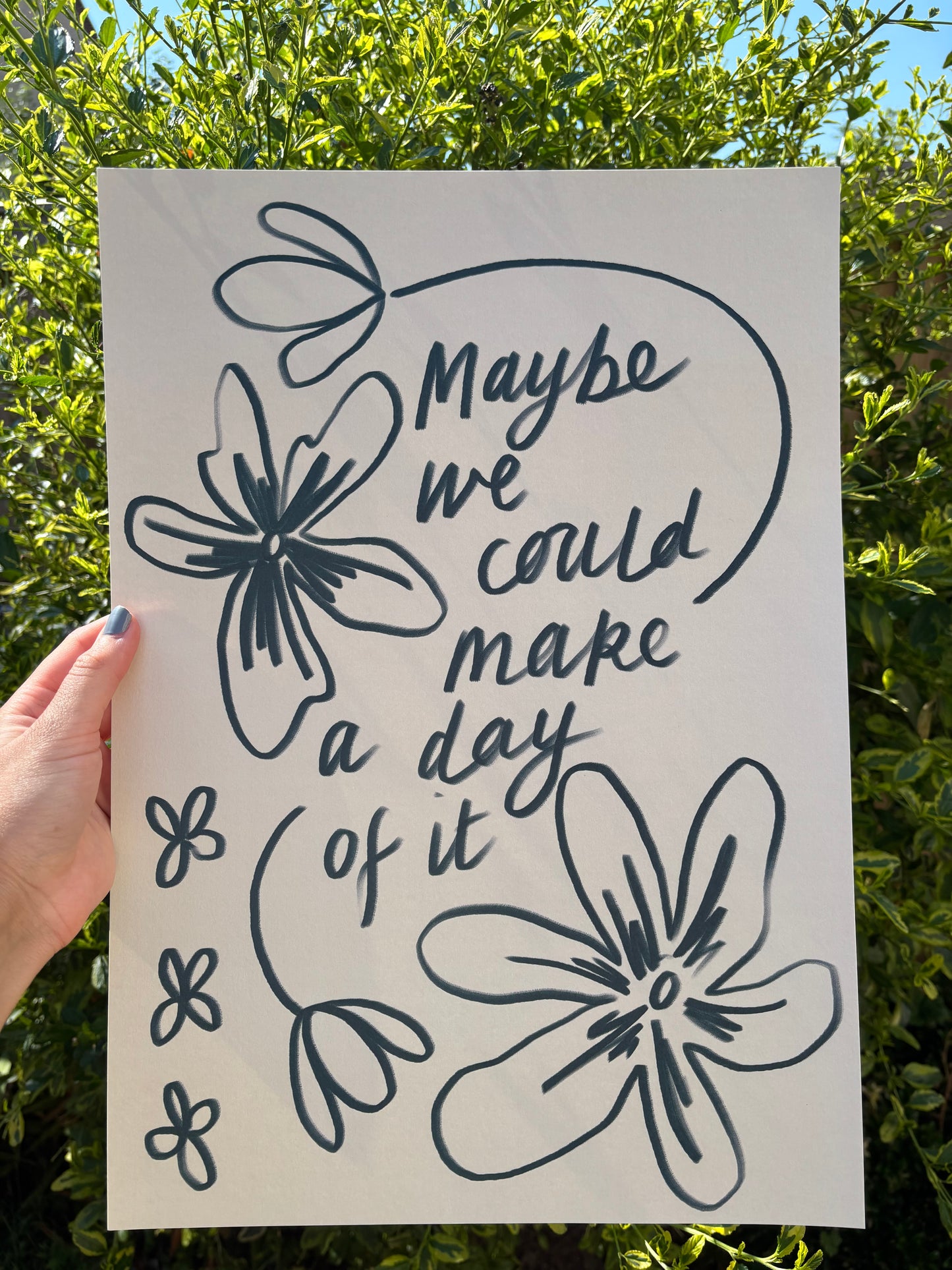 A hand holds an A3 art print outdoors against green foliage. The print features dark blue hand-drawn floral outlines and the phrase “Maybe we could make a day of it” in a script font.