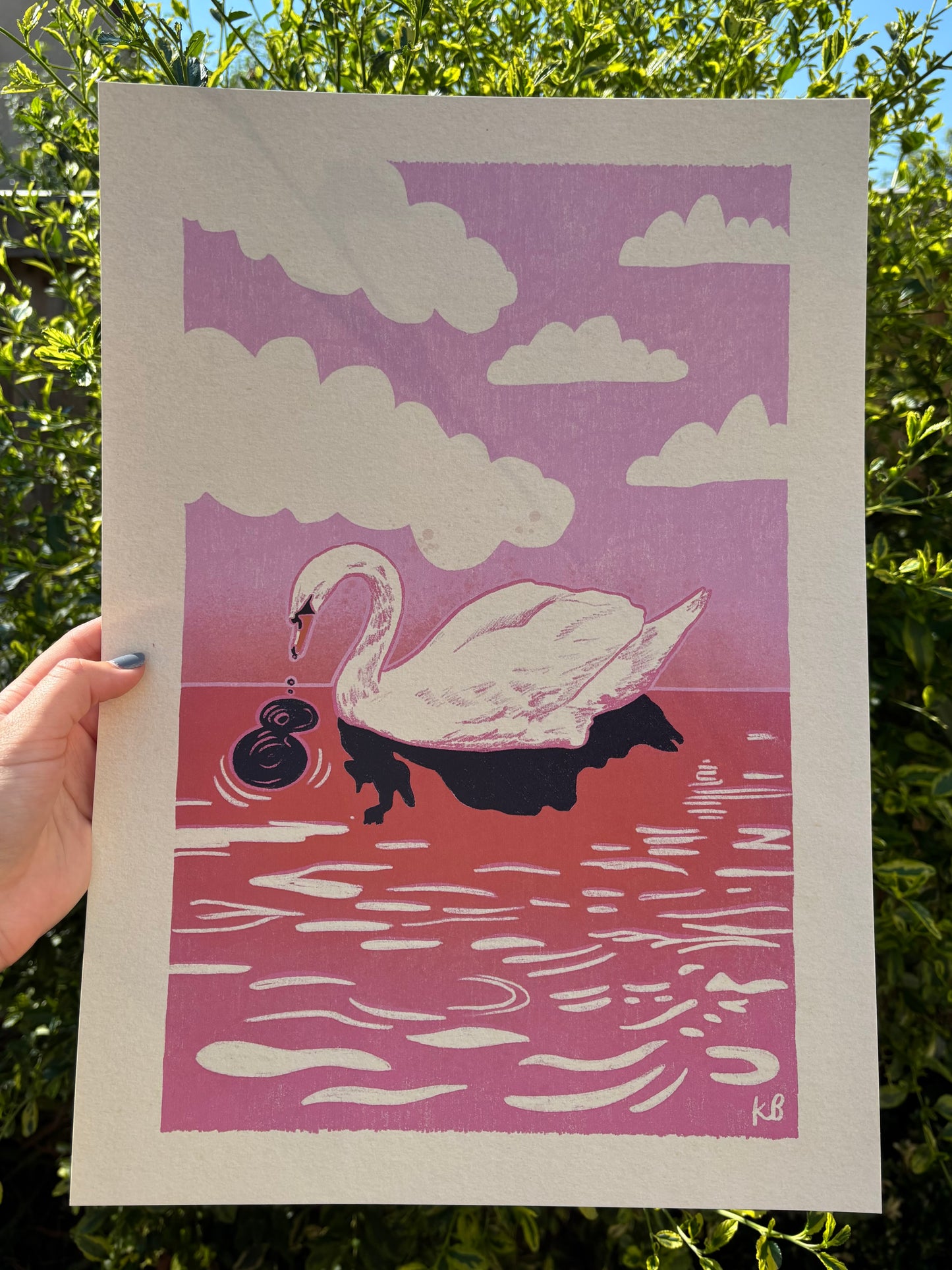 A hand holds an A3 sized art print featuring a stylised white swan on pink and red water under a pink sky with white clouds. The print is photographed outdoors against green foliage.
