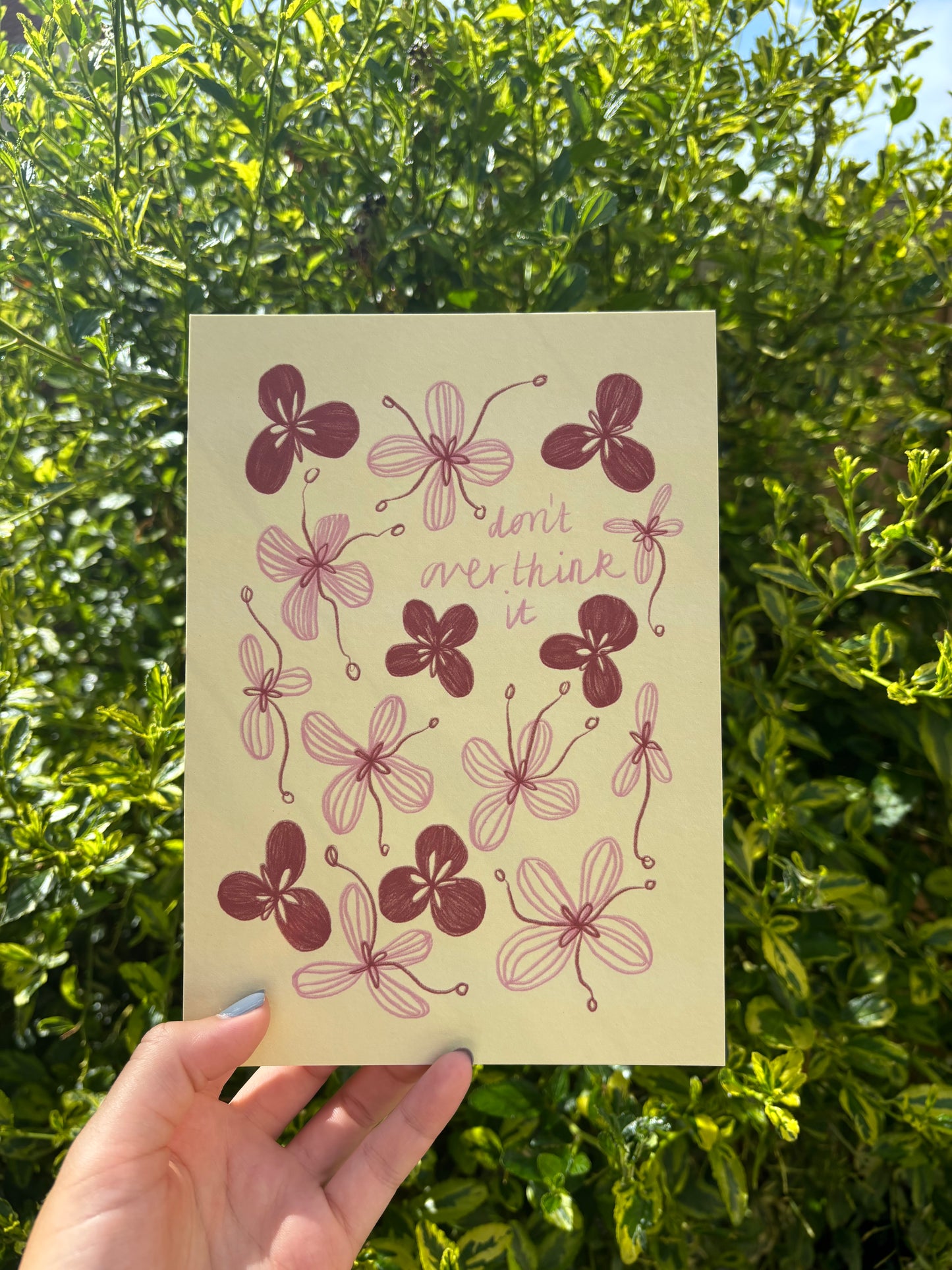 A hand holds a pale yellow art print featuring illustrated flowers in red and pink, with the text 'don't overthink it' against a background of green outdoor foliage.
