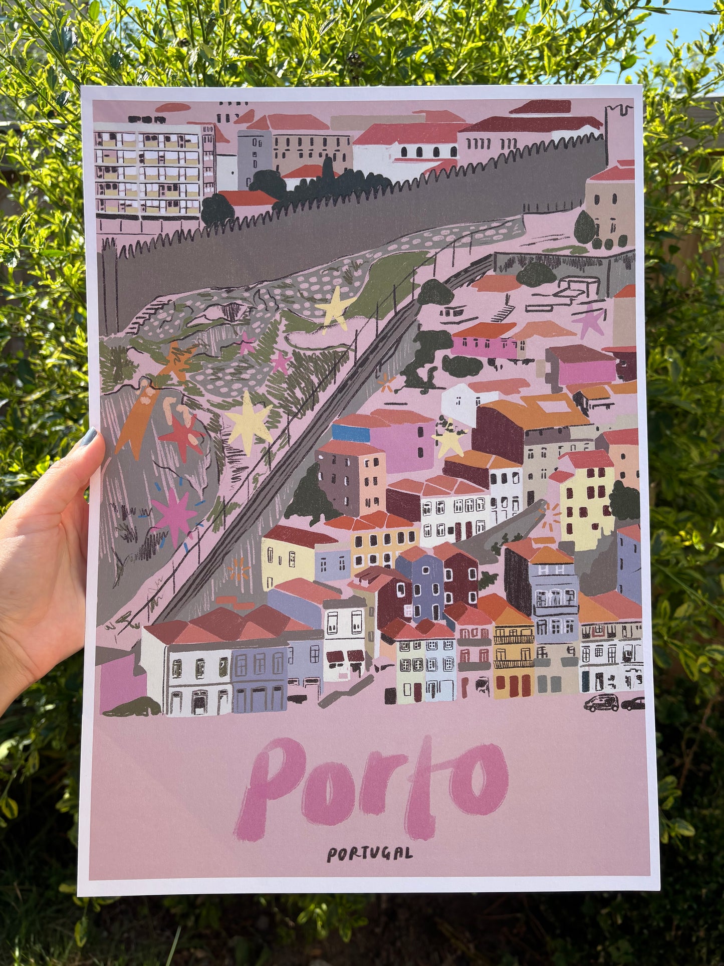 A hand holds a printed art poster featuring a stylised illustration of Porto, Portugal, with the word 'Porto' in pink on a light pink background. The artwork contains many pastel and earthy colours.