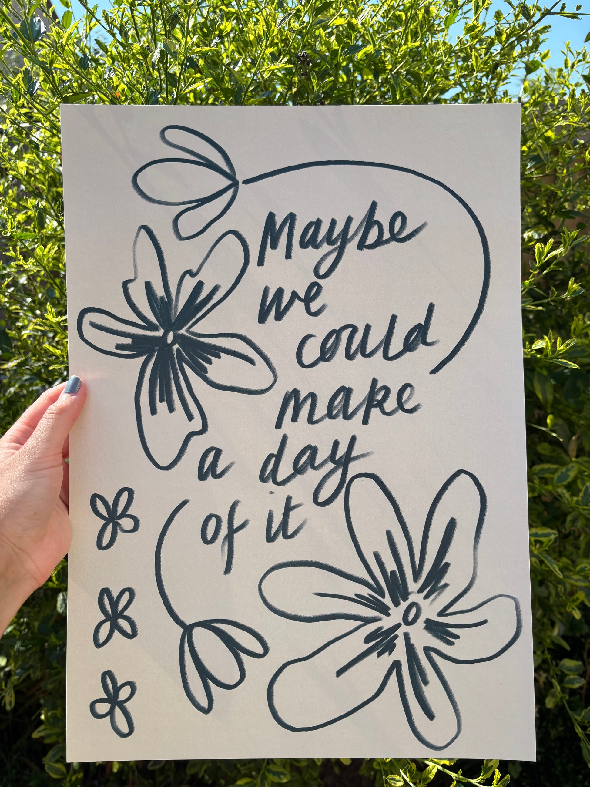 A hand holds an A3 art print outdoors against green foliage. The print features dark blue  hand-drawn floral outlines and the phrase “Maybe we could make a day of it” in a script font.