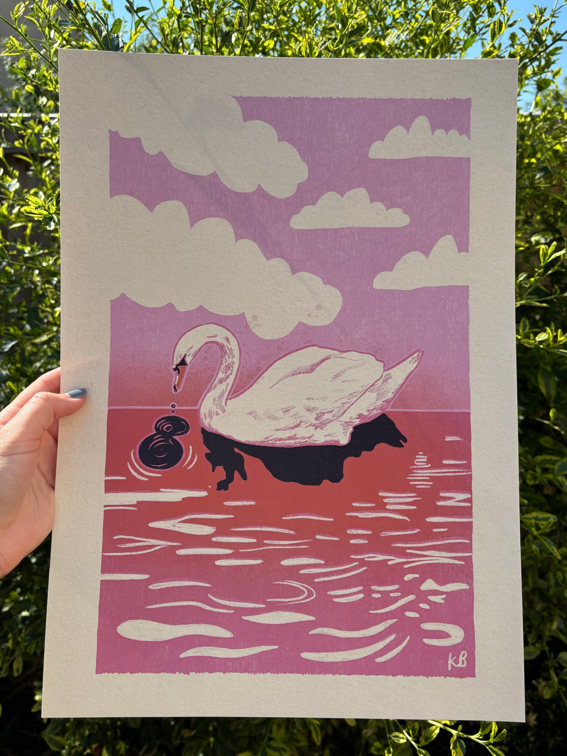 A hand holds an A3 sized art print featuring a stylised white swan on pink and red water under a pink  sky with white clouds. The print is photographed outdoors against green foliage.