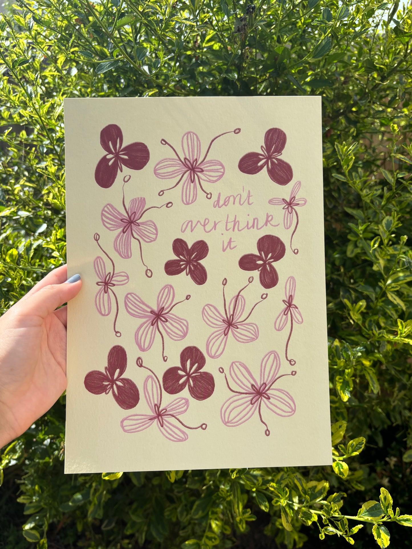 A hand holds a pale yellow A4 art print featuring pink and red flower  illustrations and the handwritten quote 'don't overthink it' against a background of green garden foliage.