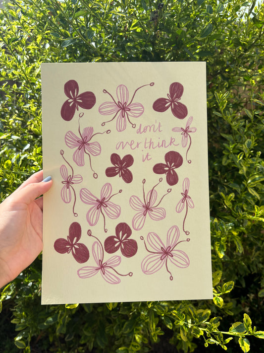 A hand holds a pale yellow A4 art print featuring pink and red flower  illustrations and the handwritten quote 'don't overthink it' against a background of green garden foliage.