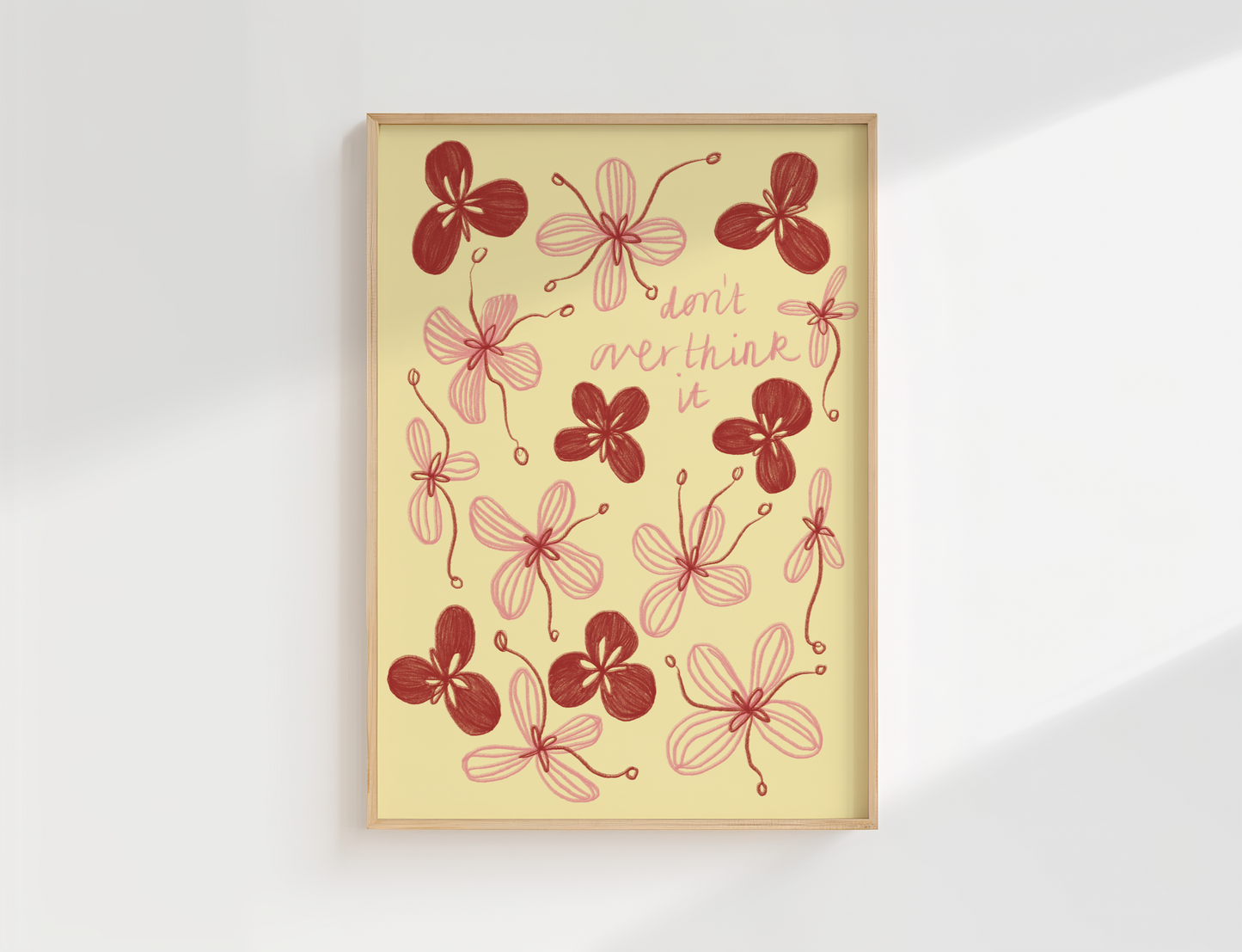 A framed art print featuring red and pink floral illustrations with the handwritten text 'don't overthink it' on a pale yellow background, displayed on a white wall with soft shadows.