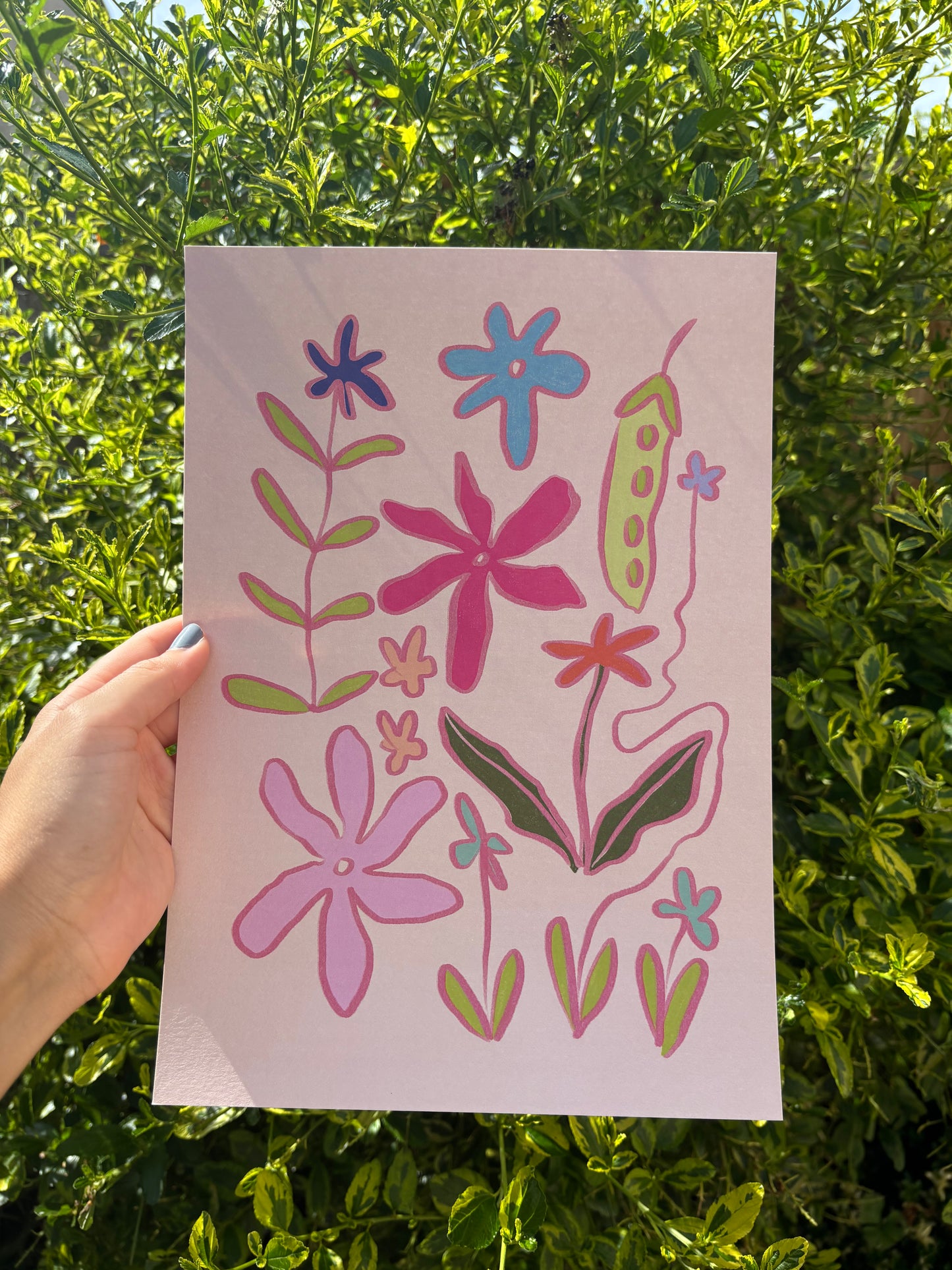 A hand holds an A4 pink art print featuring colourful hand-drawn flowers and leaves, set against a background of green outdoor foliage.