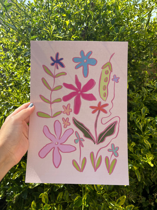 A hand holds an A4 pink art print featuring colourful hand-drawn flowers and leaves, set against a background of green outdoor foliage.