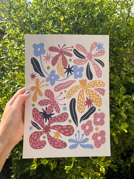 A hand holds a beige art print featuring a colourful folk-art floral pattern with pink, orange, and blue flowers, set against a background of green outdoor foliage.