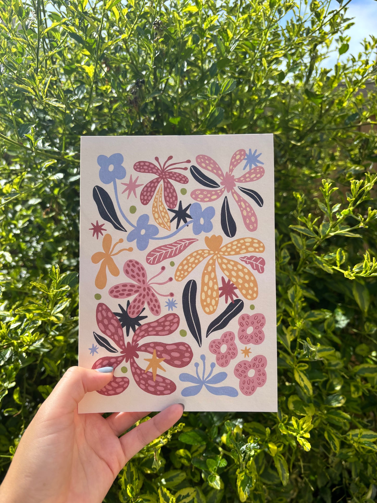 A hand holds an A5 beige art print featuring a colourful folk-art floral pattern with pink, orange, and blue flowers, set against a background of green outdoor foliage.