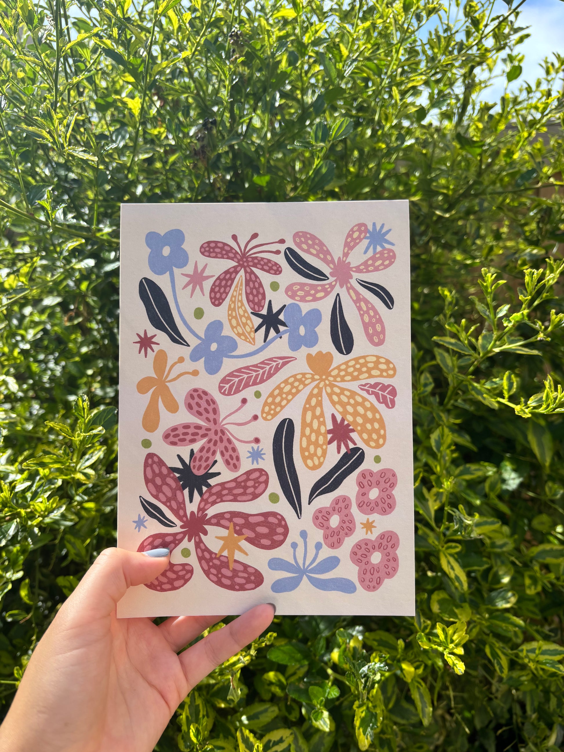 A hand holds an A5 beige art print featuring a colourful folk-art floral pattern with pink, orange, and blue flowers, set against a background of green outdoor foliage.