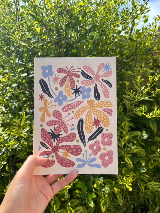 A hand holds an A5 beige art print featuring a colourful folk-art floral pattern with pink, orange, and blue flowers, set against a background of green outdoor foliage.