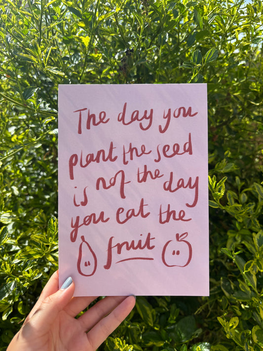 A hand holds a light pink A5 art print with red handwritten text that reads, 'The day you plant the seed is not the day you eat the fruit,' against a background of green leafy bushes.