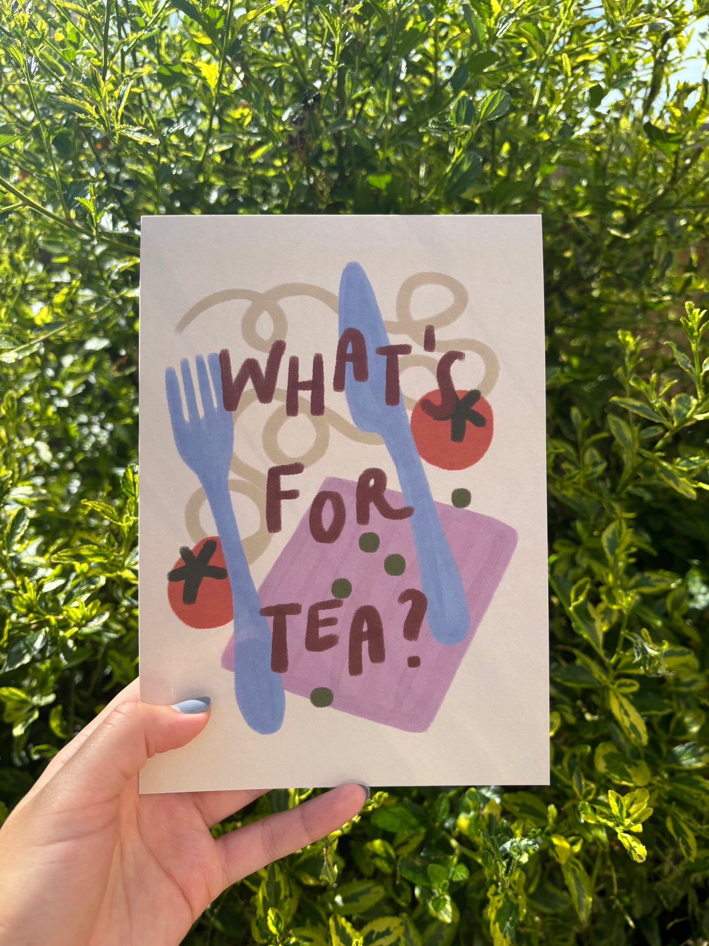 A hand holds an A5 art print in front of dense green foliage. The card features a hand-painted style illustration with the text 'WHAT'S FOR TEA?' in dark brown lettering over blue cutlery, red tomatoes, green peas, and a purple napkin.