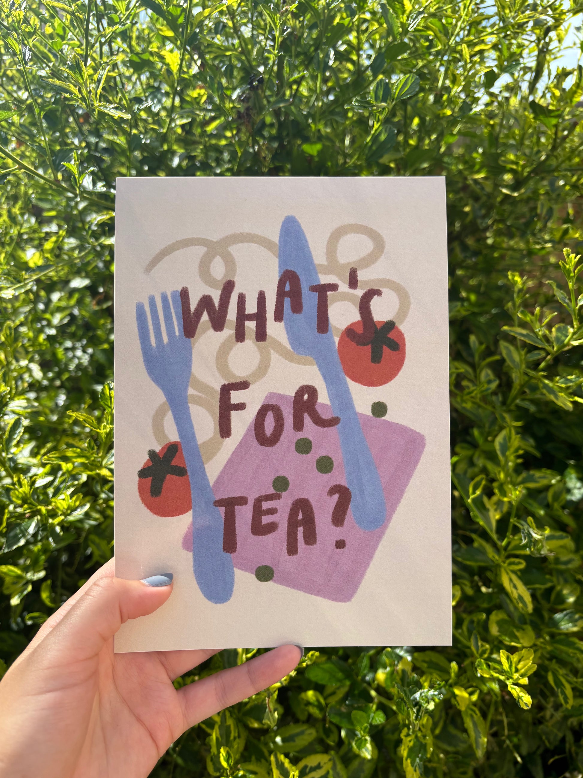 A hand holds an A5 art print in front of dense green foliage. The card features a hand-painted style illustration with the text 'WHAT'S FOR TEA?' in dark brown lettering over blue cutlery, red tomatoes, green peas, and a purple napkin.