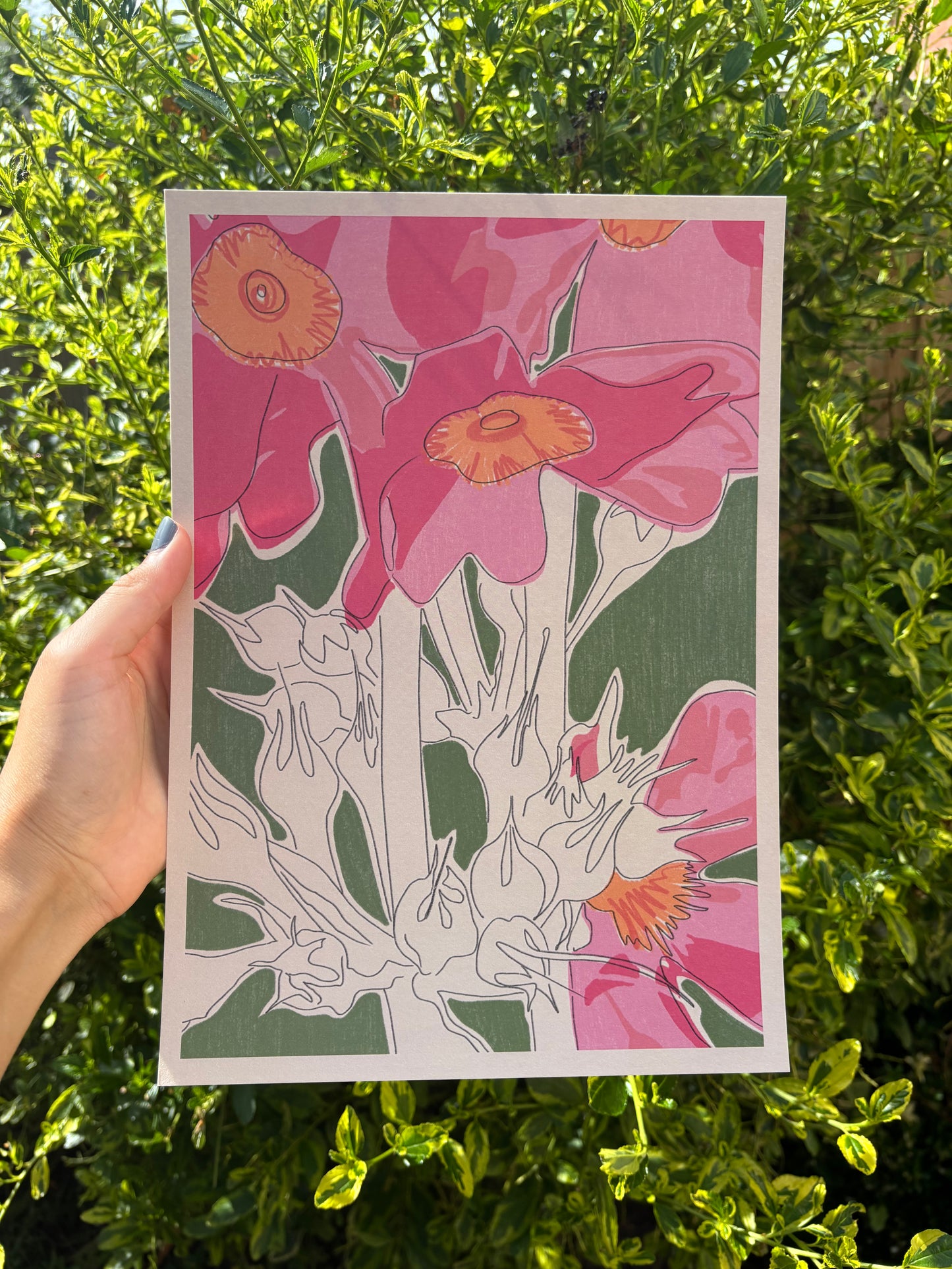 A hand holds an A4 sized art print featuring stylised pink flowers with orange centres and white line-art stems against a green background, photographed outdoors in front of dense green foliage.
