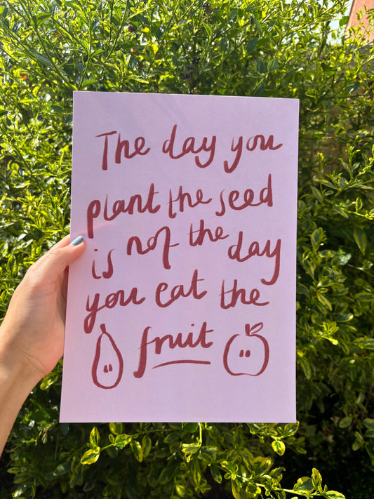 A hand holds a light pink art print featuring a hand-lettered quote ‘the day you plant the seed is not the day you eat the fruit’ set against a background of green garden foliage.