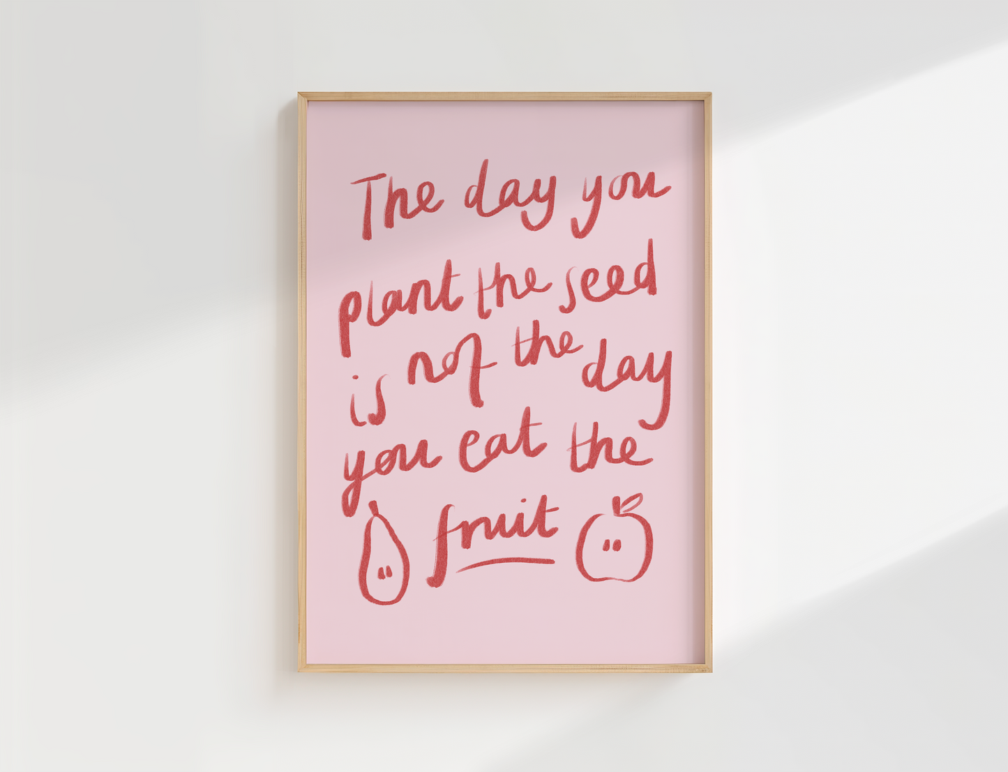 A framed wall art print with a light pink background and red handwritten text reading, 'The day you plant the seed is not the day you eat the fruit,' plus simple fruit illustrations. The frame is light wood, shown hanging against a white wall with soft diagonal shadows.